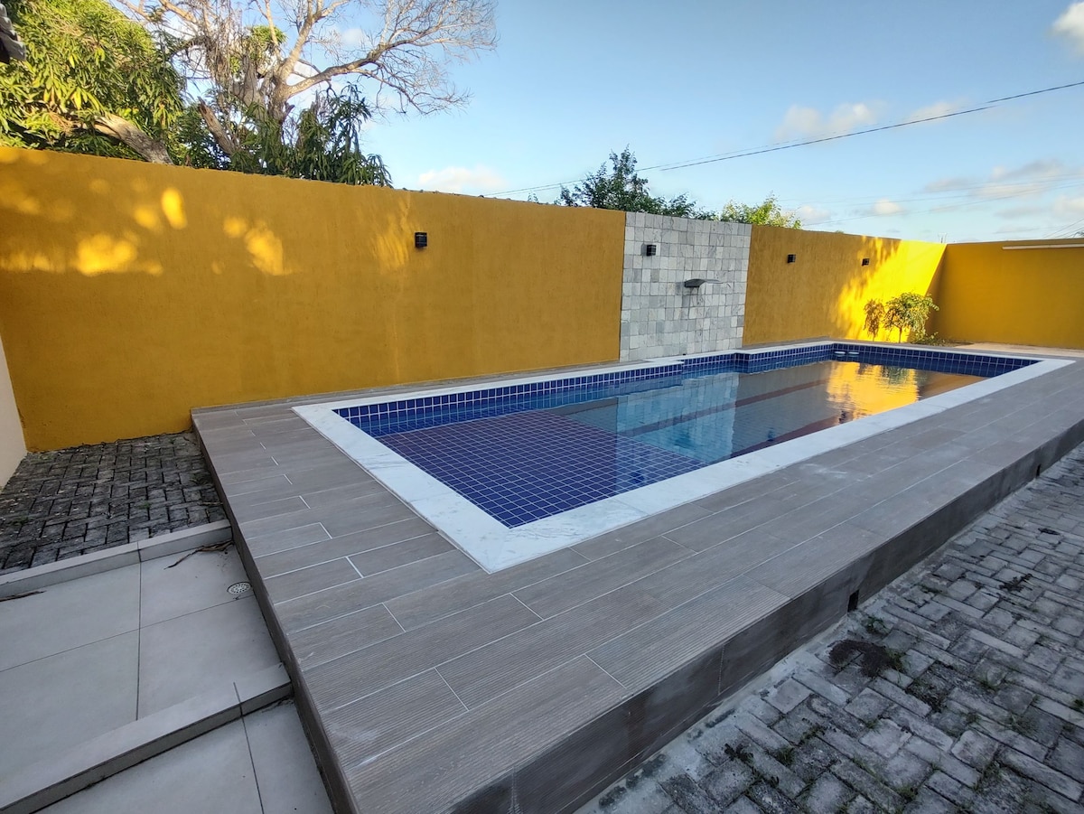 A well-maintained pool area features a rectangular swimming pool surrounded by a wooden deck. The pool's blue tiles contrast with a vibrant yellow wall in the background. Lush greenery is visible near the edge, adding to the serene environment.