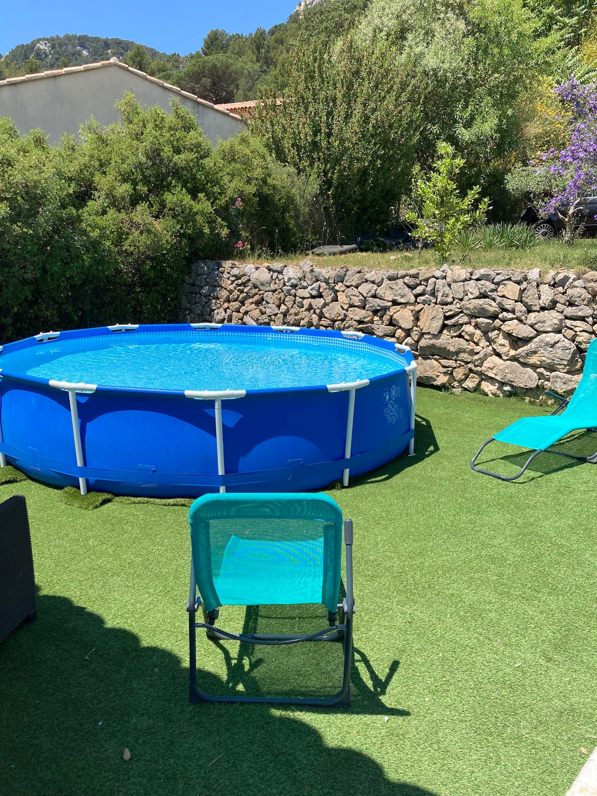 A private above-ground pool is surrounded by a neatly kept garden, featuring artificial grass. A single folding chair is positioned nearby, with two turquoise lounge chairs also present. Lush greenery and blooming plants provide a tranquil backdrop.