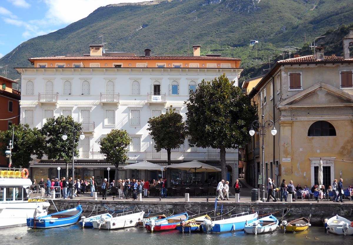 A view of a vibrant waterfront scene features a charming building with a balcony, alongside a bustling promenade filled with people. Colorful boats are docked in the foreground, while trees provide greenery along the waterfront. The backdrop showcases the mountains rising above the town.
