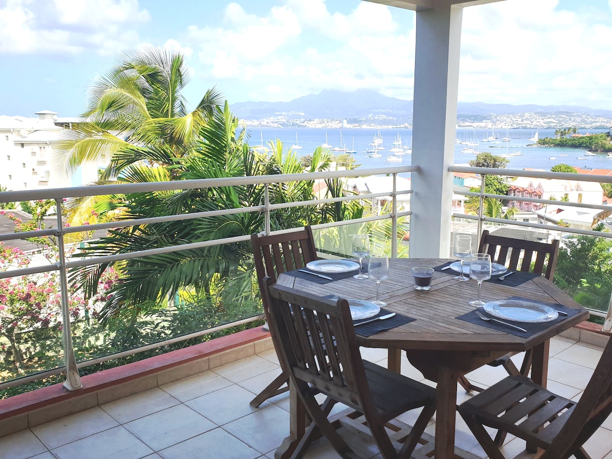 A terrace is showcased, featuring a round wooden table set for four, surrounded by matching chairs. Lush greenery and distant views of the water and mountains create a serene backdrop. The space is designed for outdoor dining and relaxation.