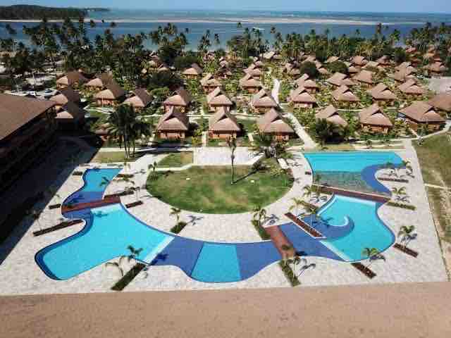 An aerial view captures the resort's spacious grounds, showcasing two swimming pools with varying shapes and a central green area. Thatched-roof accommodations are arranged around the pools, surrounded by palm trees and a serene ocean in the background.