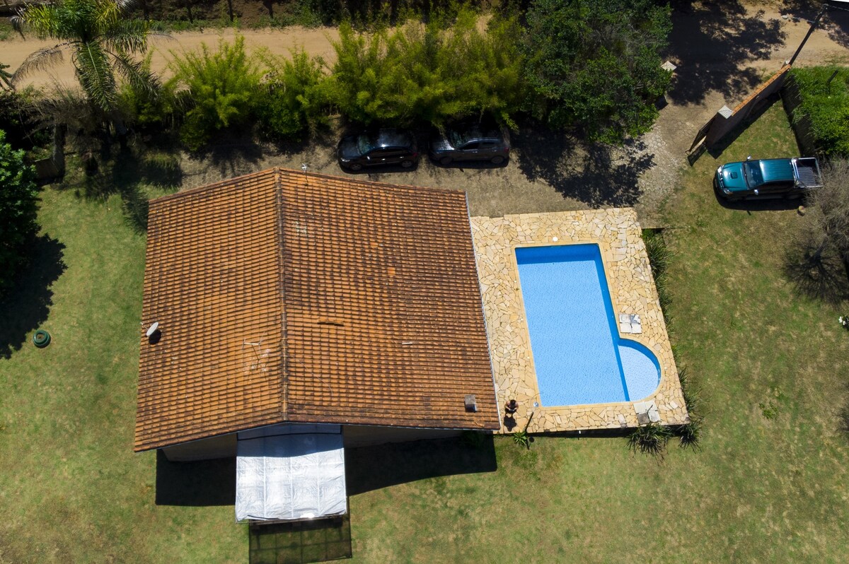 An aerial view captures a rustic house with a terracotta roof adjacent to a rectangular swimming pool. Surrounding greenery includes trees and shrubs, and pathways approach the house and pool area. Two vehicles are parked along a gravel drive.
