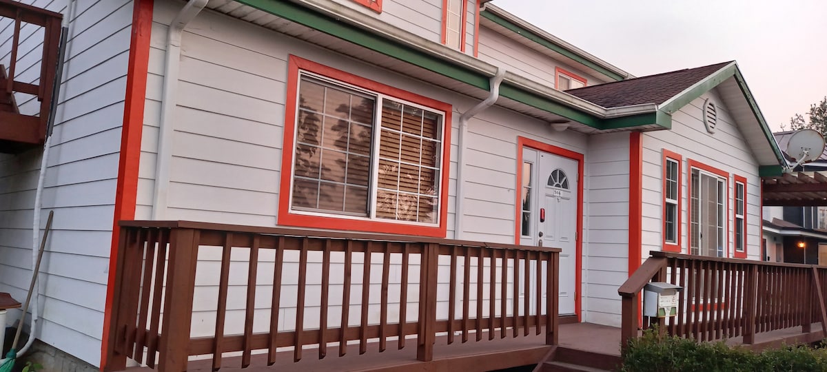 A two-story building is depicted, featuring white exterior walls with vibrant orange trim. A wooden porch extends in front, with a railing surrounding the entryway. Several windows allow natural light to illuminate the interior, showcasing a welcoming facade.