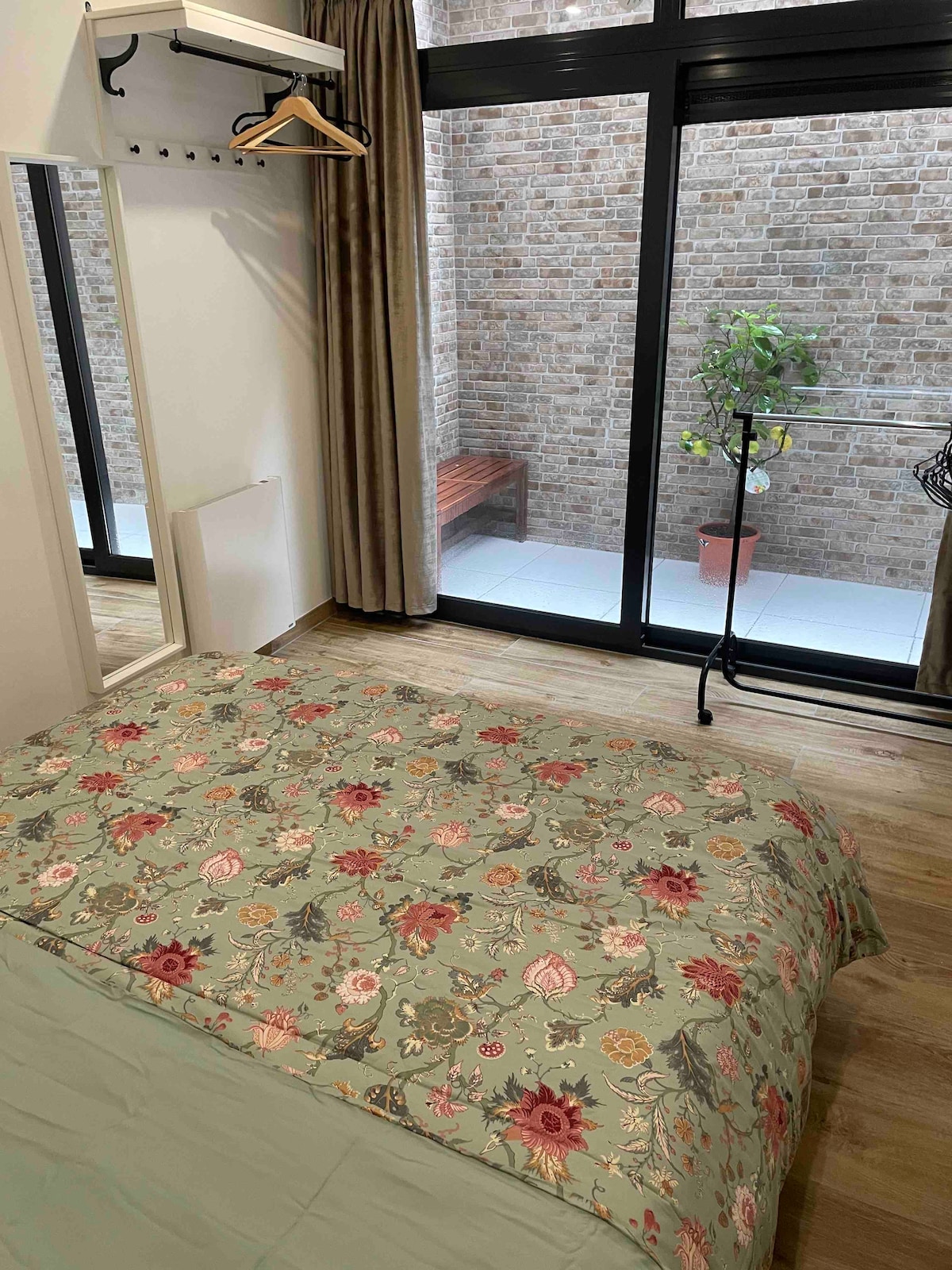 A spacious sleeping area features a large bed adorned with a floral-patterned cover. Natural light enters through generous glass doors, revealing a small outdoor space with a potted plant and a wooden bench. A mirror and clothing rack are positioned conveniently nearby.
