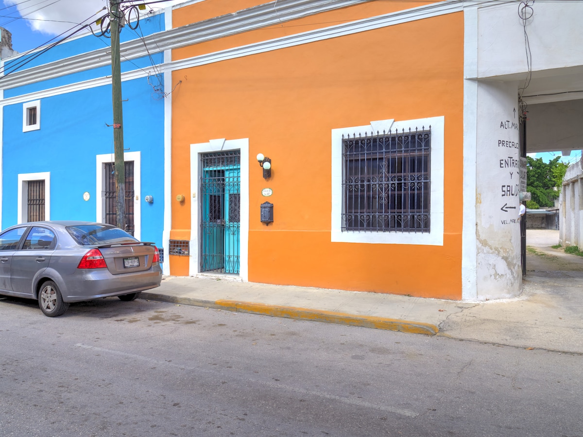 A vibrant colonial house is portrayed, featuring bright blue and orange exterior walls. The entrance is highlighted by a turquoise door and decorative iron fencing. Windows with iron bars provide views into the house. A silver car is parked on the street in front.