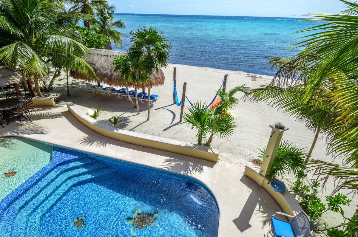 A large heated pool curvily frames the foreground, offering a striking view of the sandy beach and tranquil sea beyond. Palm trees and hammocks enhance the beachside setting, while lounge chairs are arranged on the sand, inviting relaxation under the sun.