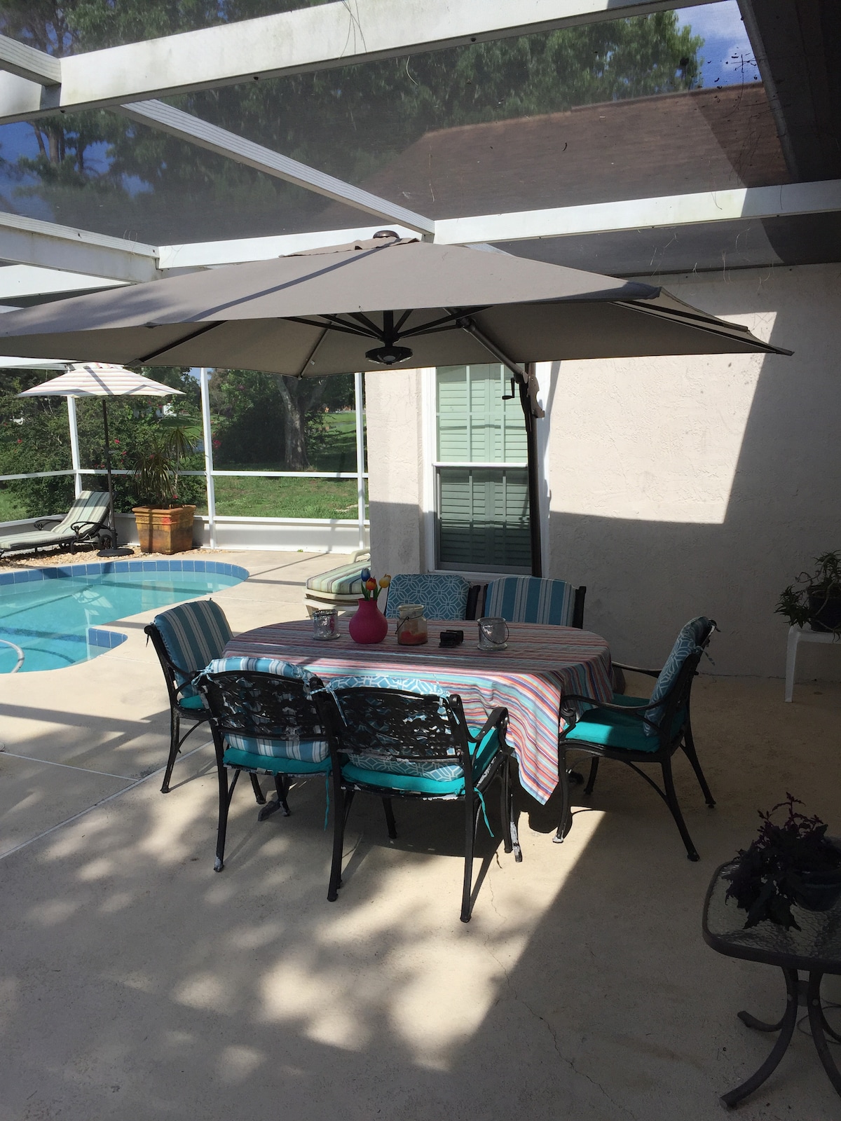 A shaded outdoor patio area features a round table surrounded by six stylish chairs, with a colorful striped tablecloth. A large umbrella provides shade over the table, while the nearby pool glimmers in the sunlight, framed by a tranquil green landscape.