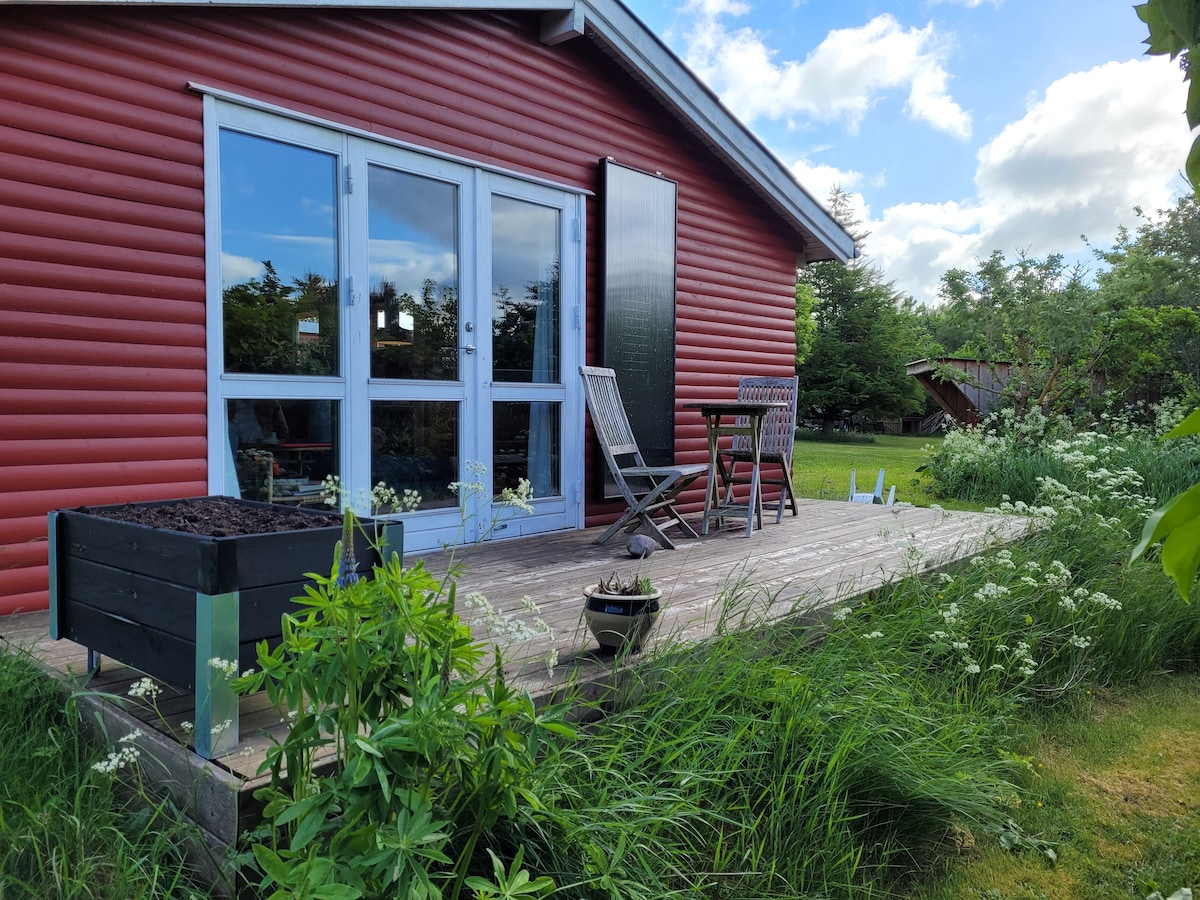 A red wooden house is seen with large glass doors opening to a spacious deck. An outdoor table with two chairs is set on the deck, surrounded by lush green grass and plants. A planter box is placed nearby, enhancing the natural surroundings.