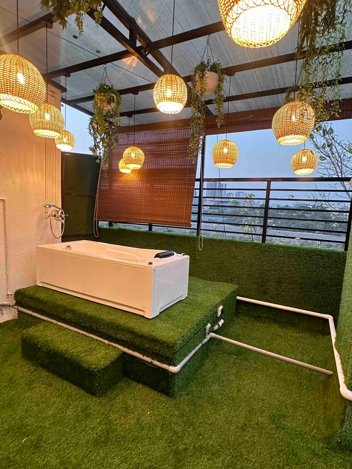 A private terrace area features a raised jacuzzi surrounded by lush green turf. Ambient lighting from woven pendant lamps hangs above, adding a soft glow to the space. The atmosphere is enhanced by greenery along the walls, providing a serene setting for relaxation.