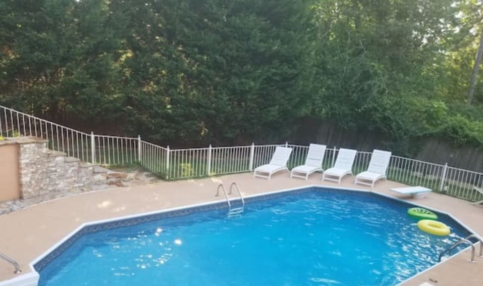 A large pool is surrounded by a spacious deck and features steps leading from the patio. Several white lounge chairs are positioned along the edge. A safety fence encloses the area, with greenery providing a natural backdrop.