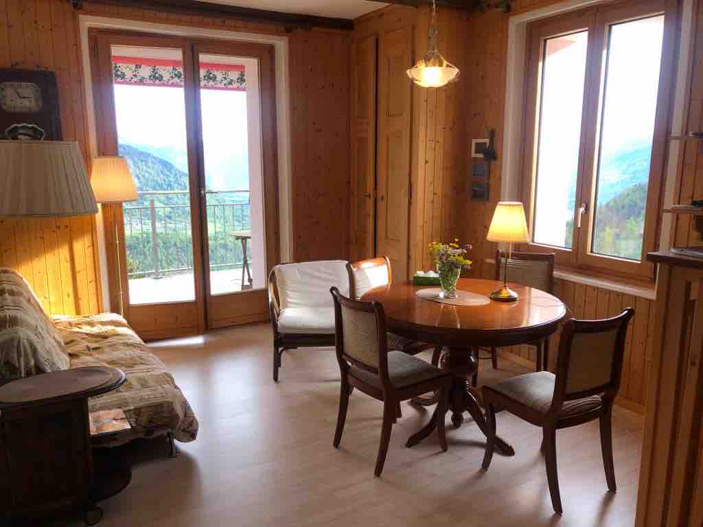 A cozy living and dining area is featured, showcasing a wooden table surrounded by four chairs. A comfortable sofa sits beside a window, which offers access to a balcony. Light is provided by lamps and a ceiling fixture, enhancing the natural wooden decor.