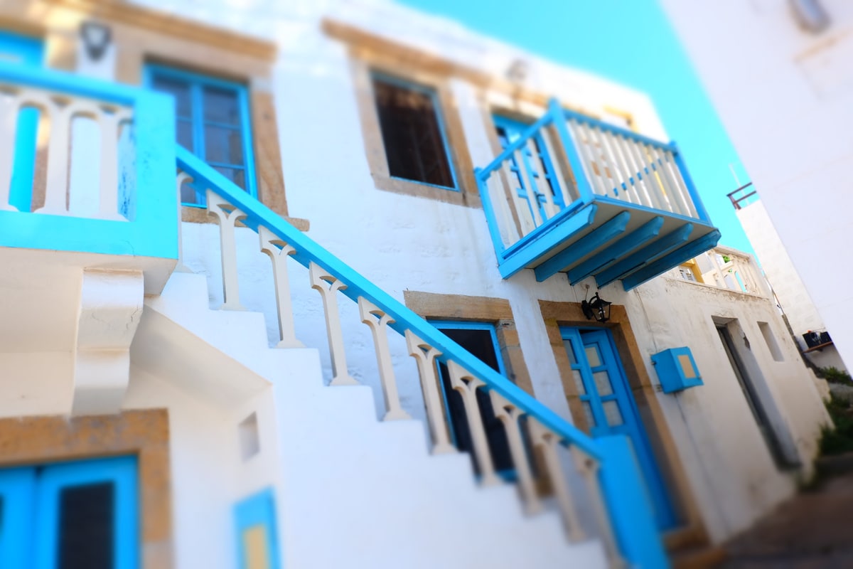 The exterior of a charming two-story house is presented, featuring white walls complemented by vibrant blue accents. A staircase leads to a second-floor balcony, while a door is visible at the ground level. The surrounding area is well-maintained, suggesting comfort and accessibility.