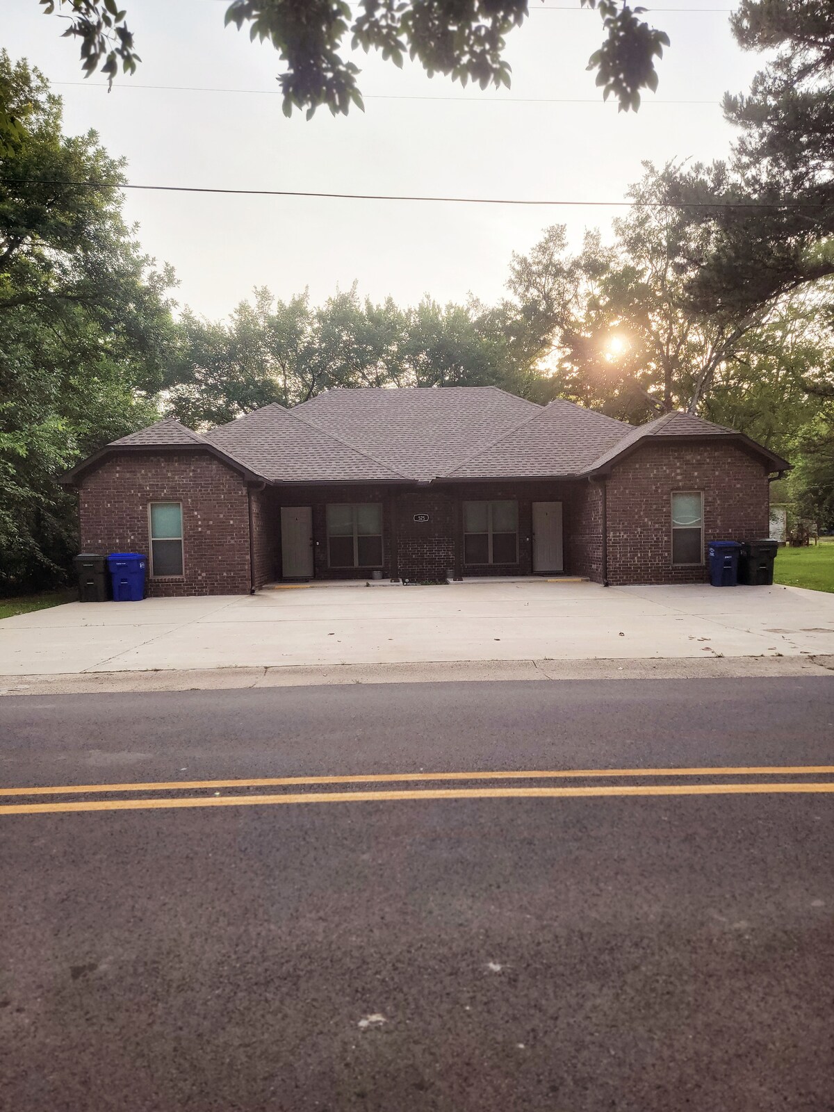 A brick duplex is showcased in the image, featuring a symmetrical facade with a sloped roof. Two entryways are visible, flanked by large windows. A paved driveway extends in front, with blue recycling bins placed near the exterior. Trees line the background, providing a natural setting.
