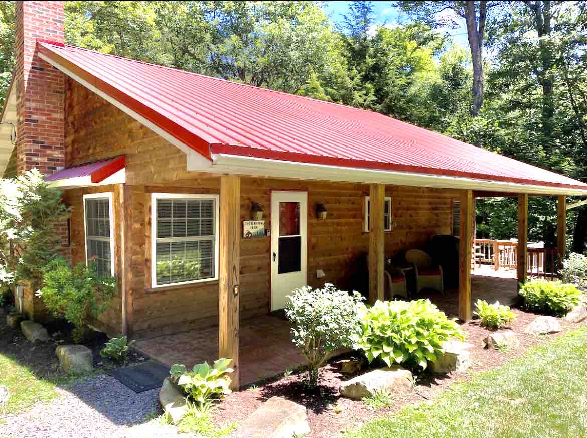 The cabin features a charming exterior with wood siding and a bright red metal roof. A welcoming porch is supported by wooden pillars, with seating positioned for relaxation. Lush greenery surrounds the entrance, complementing the natural setting.