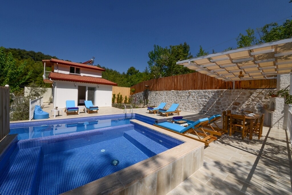 A private pool area with a clear blue surface is surrounded by sun loungers and wooden chairs. A shaded dining table sits nearby, and a stone wall adds texture to the scene. The villa features a red-tiled roof and is framed by lush greenery.