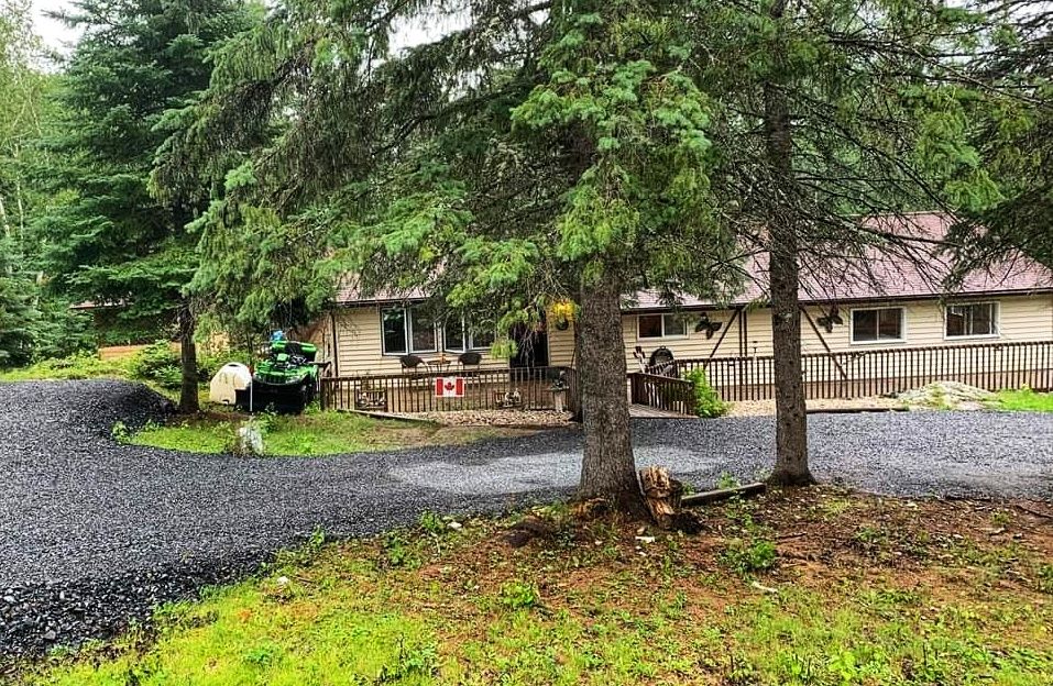 A spacious cabin is nestled within a wooded area, surrounded by tall trees. A gravel driveway leads up to the entrance, where a covered porch offers seating options. The front deck extends along the cabin, providing a welcoming outdoor space.