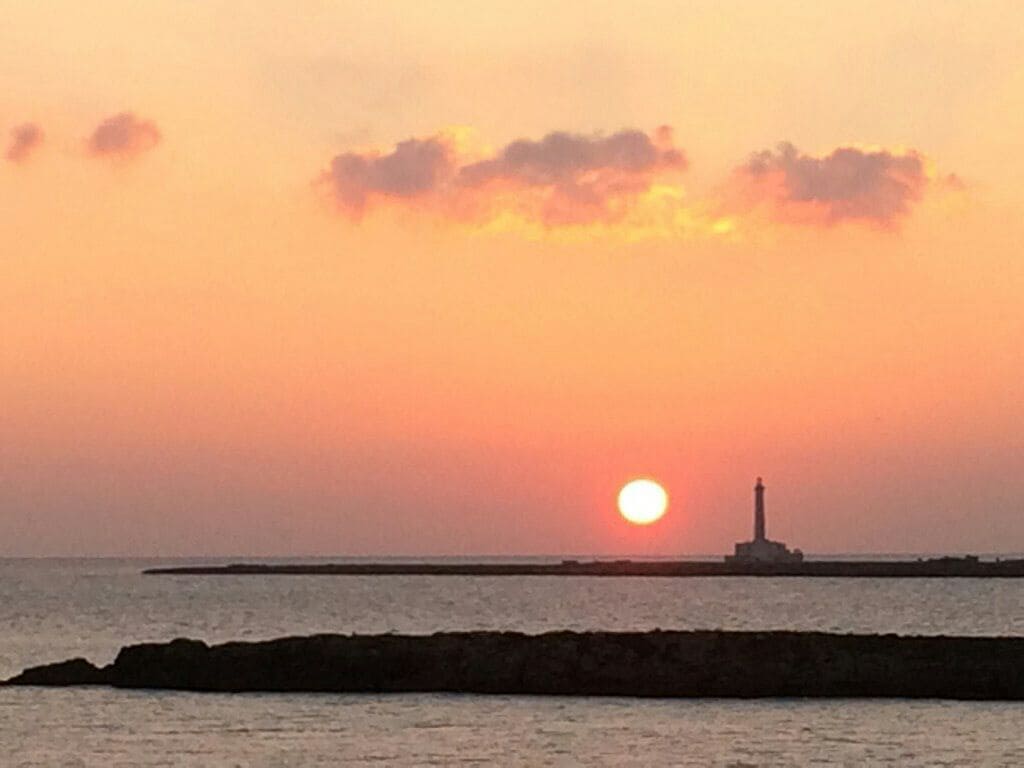 A serene sunset is depicted over the water, with the sun setting behind a lighthouse on a distant jetty. The sky is filled with soft hues of pink and orange, reflecting on the calm surface of the sea.