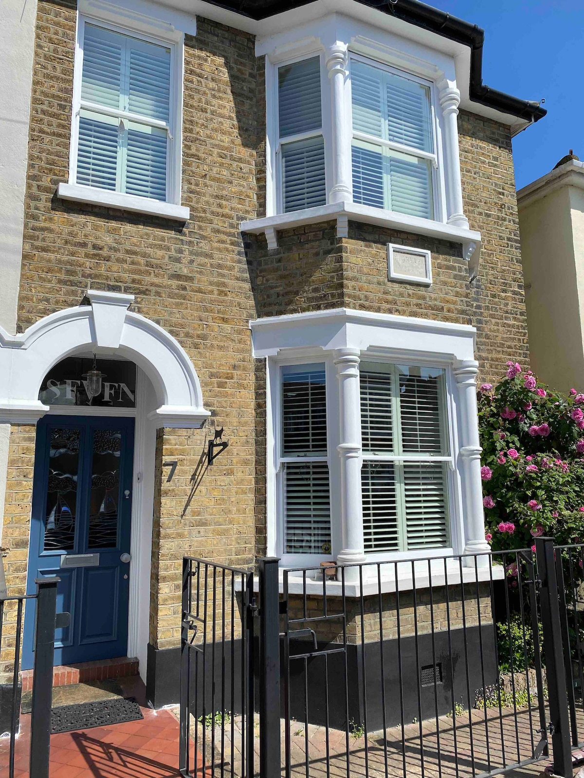 A charming front exterior of a family house is presented, featuring a distinctive brick facade with white detailing. Large windows adorned with shutters allow natural light to illuminate the entryway. A decorative gate provides access to the front path, bordered by flowering greenery.