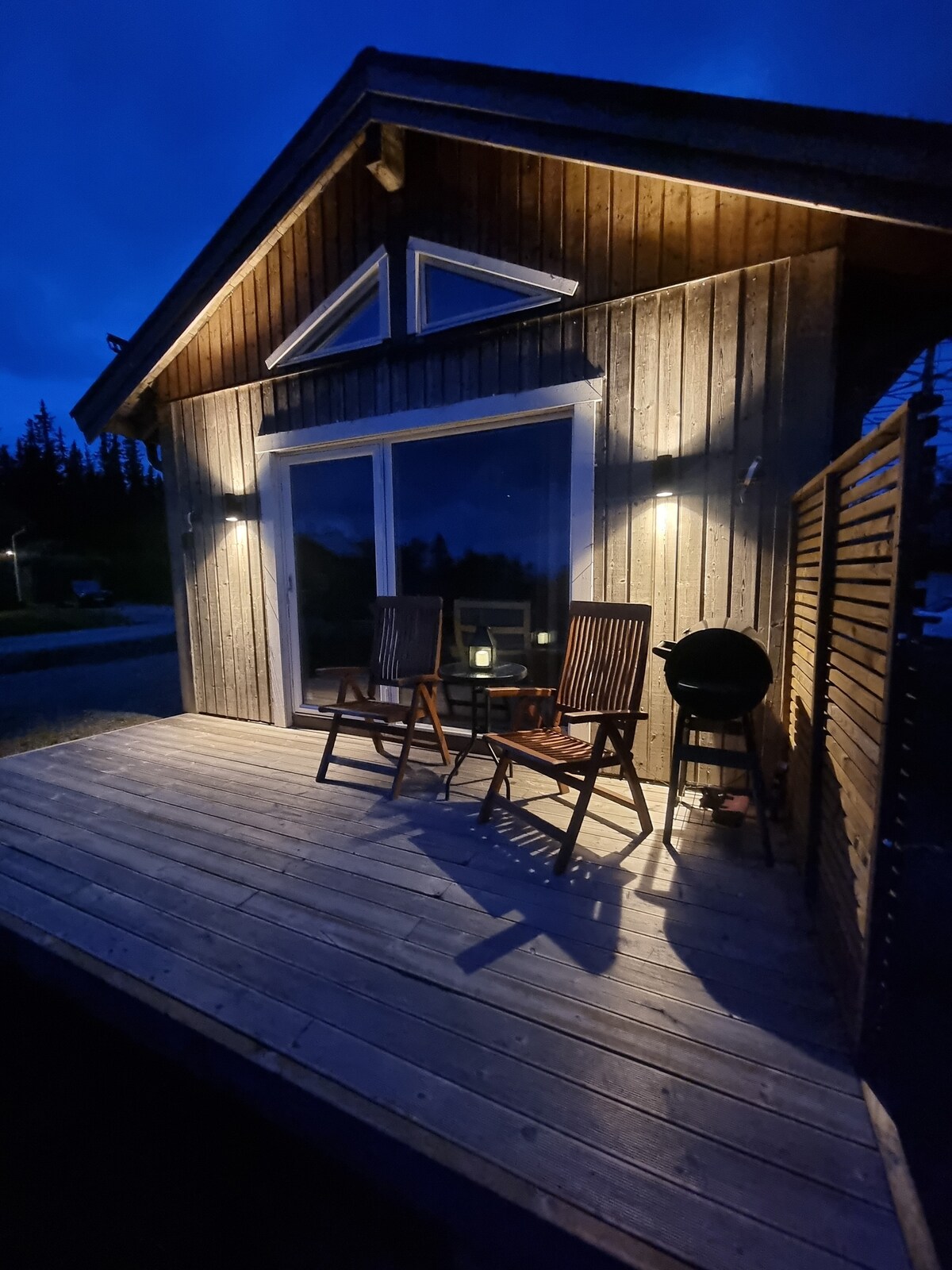 The outdoor patio showcases two wooden chairs positioned beside a grill, illuminated by soft lighting. The wood-paneled exterior extends into the night sky, reflecting a warm and welcoming ambiance in a natural setting.