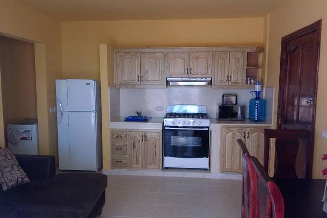 A functional kitchen space features wooden cabinetry and a gas stove. A large refrigerator stands beside a counter with small appliances. A dining area with a table and chairs complements the inviting atmosphere. Natural light illuminates the tiled floor, creating a bright environment.