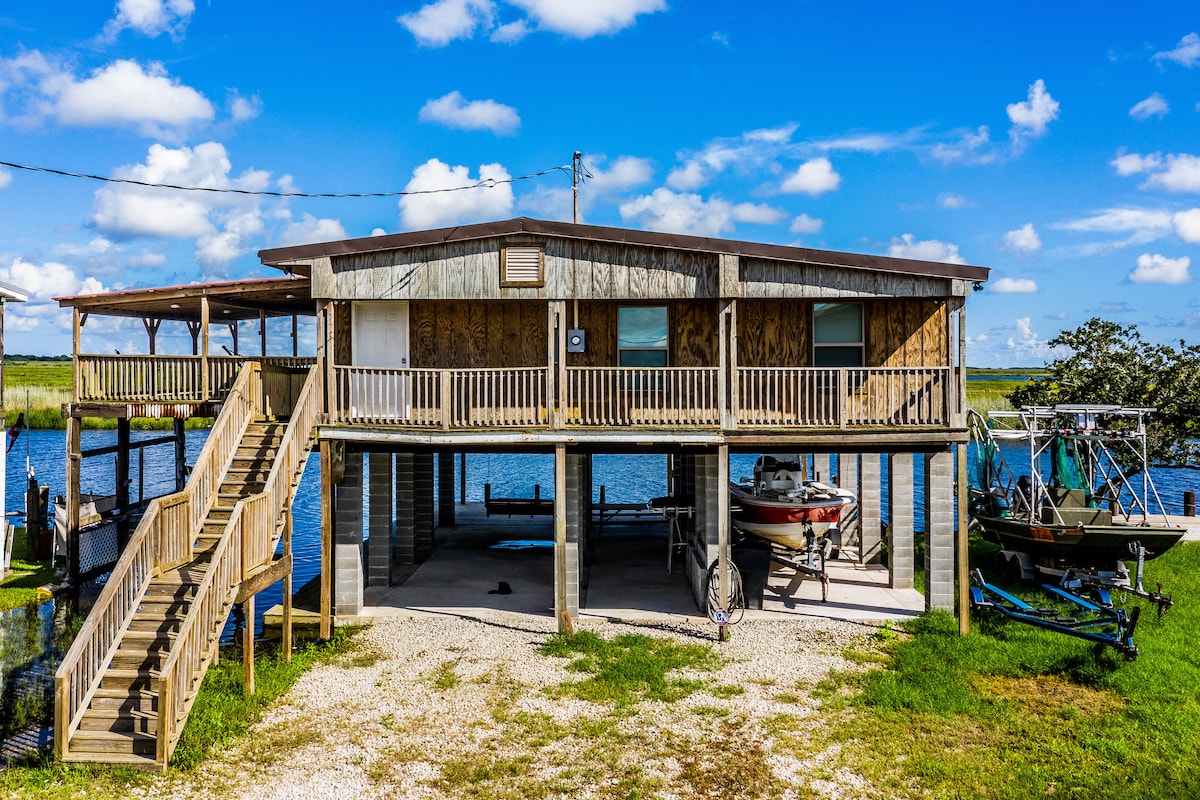 A two-story camp building is situated by the water, featuring a spacious raised deck with multiple entries. Underneath, boats are parked alongside a gravel area. The surrounding landscape includes grassy patches with a view of the bayou and the blue sky above.