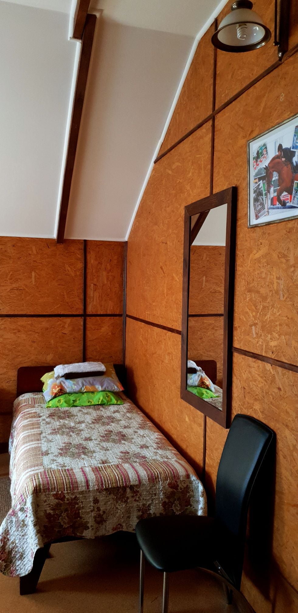 A simple bedroom features a twin bed with layered bedding and decorative pillows. A tall mirror is positioned on the wall, alongside a black chair. The walls are finished in textured wood, adding warmth to the space, while natural light illuminates the room.