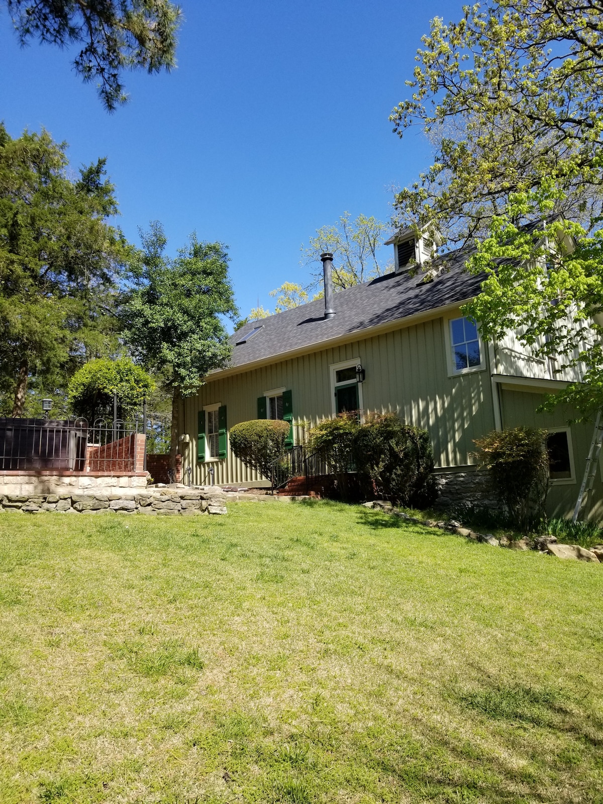 The exterior of the carriage house features a well-maintained lawn and distinct greenery. A stone border edges the yard, leading to a wooden patio area with fencing. The structure is framed by trees, and a chimney is visible on the roof, suggesting warmth inside.