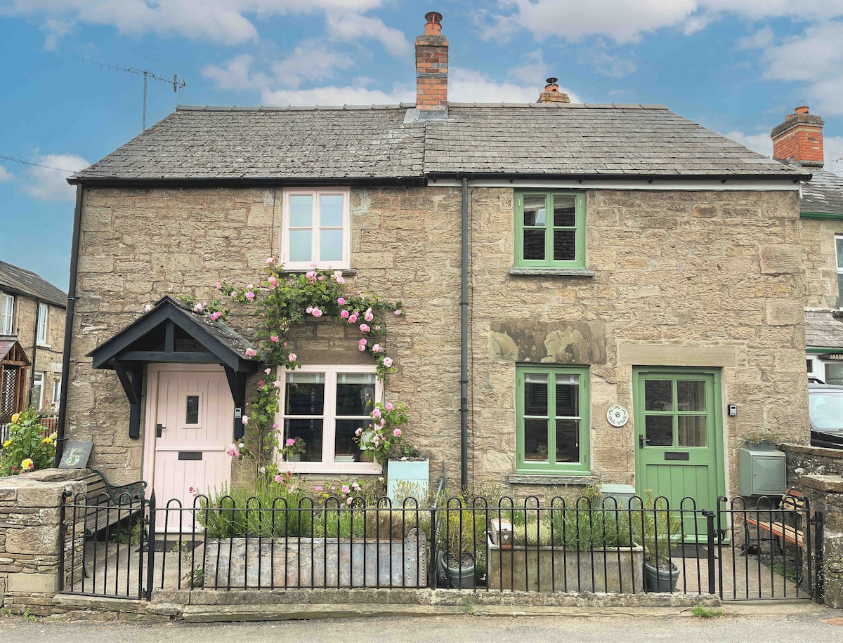 A charming stone cottage is displayed, featuring a welcoming pink front door framed by climbing flowers. Green window shutters complement the rustic exterior. A black railing surrounds the front yard, which is adorned with a small patch of grass and flowers.