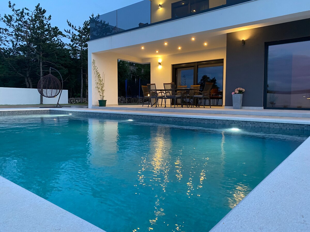 A view of the villa's swimming pool reflects soft evening lights. The pool area is complemented by a spacious terrace featuring a dining table and chairs, along with potted plants. The modern architecture of the villa showcases large windows and a covered seating area.