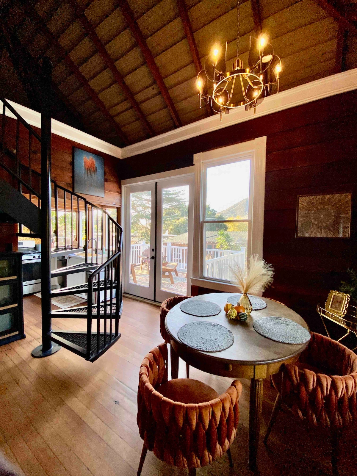 A spiral staircase is seen in a well-lit corner, leading to the upper bedrooms. A round dining table with elegantly woven chairs is positioned by large windows, offering views of the outdoor deck. Modern decor complements the rustic wooden walls and high ceiling.