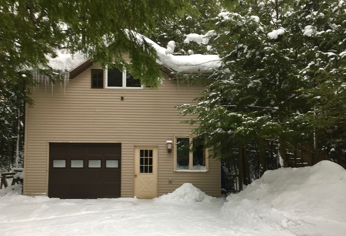 The exterior of the guest house is surrounded by snow-covered evergreen trees, showcasing a neutral siding and a large garage door. Icicles hang from the roof's edge, while an inviting front entrance is visible. Snow drifts accumulate at the base, creating a winter scene.