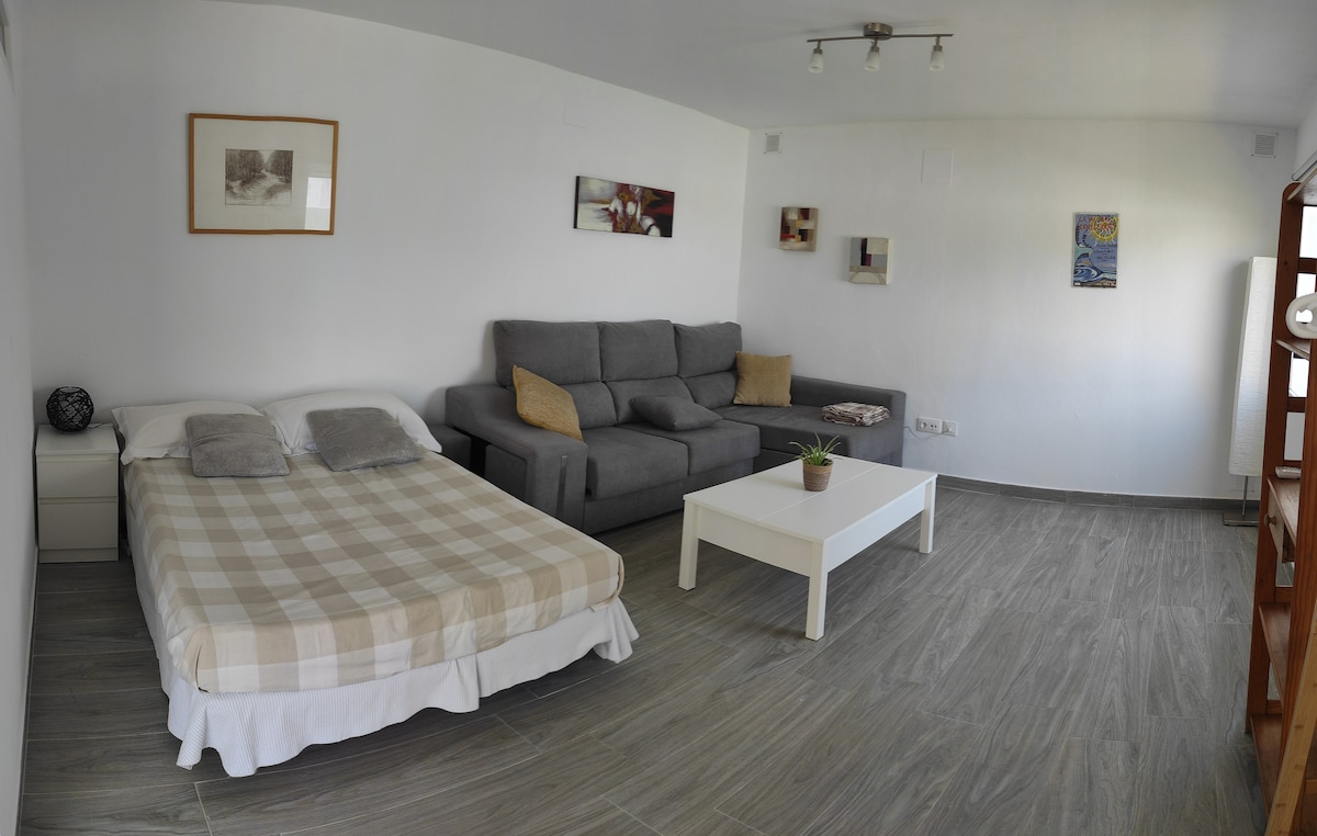 A neutral-toned room is presented with a double bed featuring a checkered blanket and soft pillows. A spacious gray sofa with chaise lounge is visible next to a white coffee table. Framed artwork decorates the walls, and the floor is tiled in light tones.