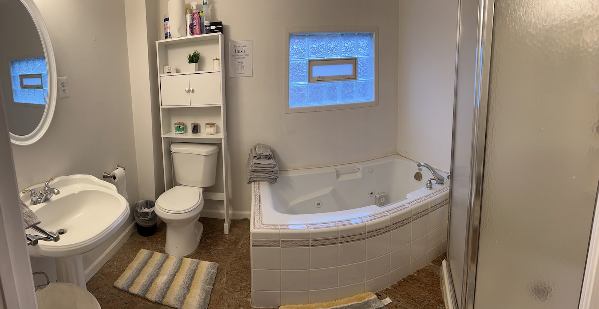 A spacious bathroom features a freestanding bathtub beside a shower stall with a glass door. A circular mirror hangs above a sink, accompanied by a shelving unit displaying toiletries. Natural light enters through a small window, illuminating soft towels arranged on the countertop.