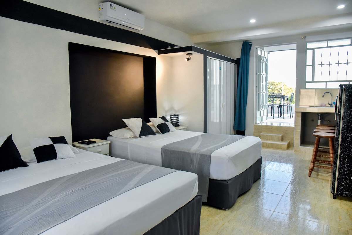 A modern room features two spacious beds with neutral bedding and decorative pillows. Natural light enters through large glass doors, leading to a private terrace. A kitchenette with bar stools is visible in the corner, complemented by a shower area separated by a glass partition.