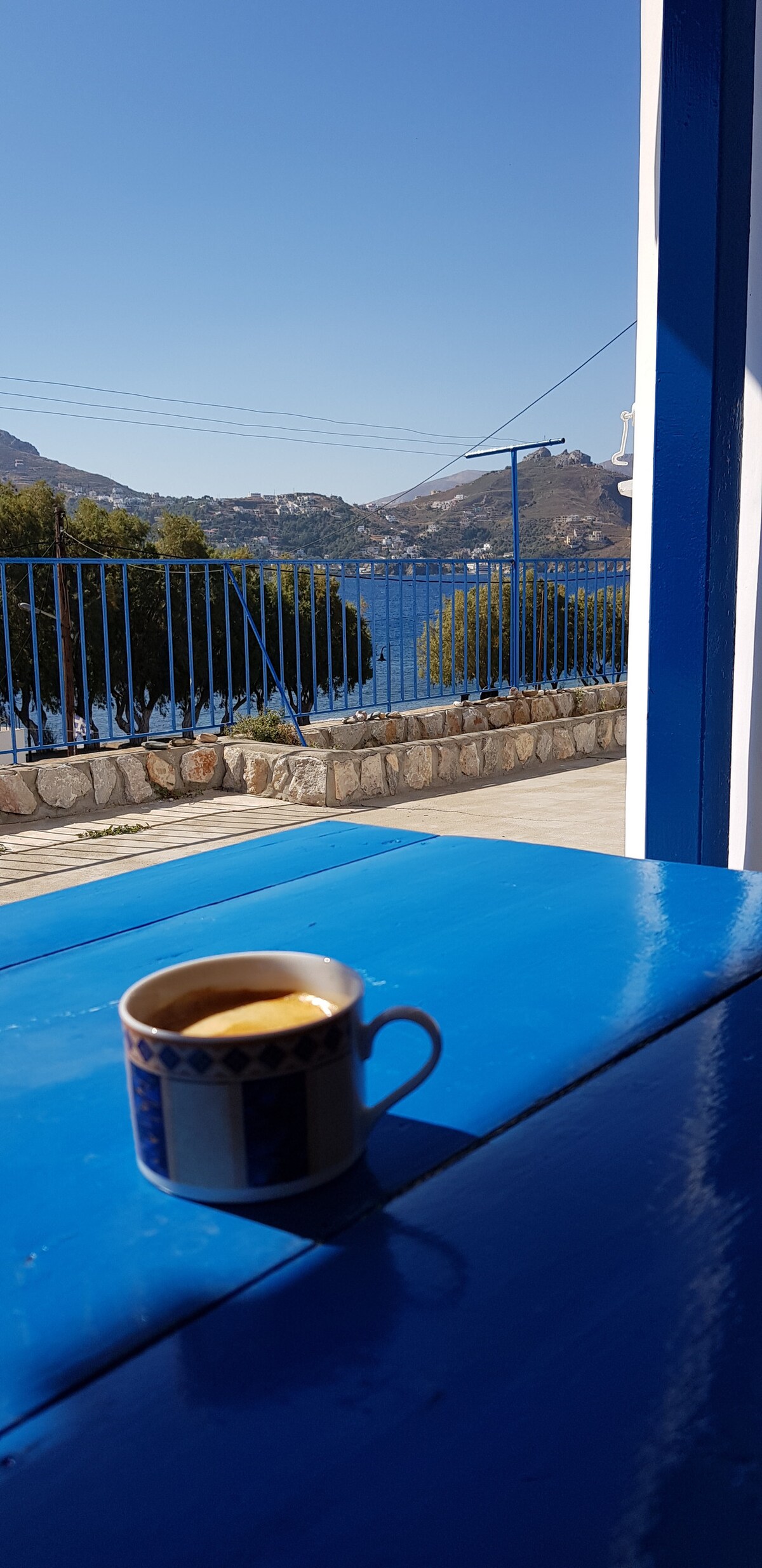 A blue table is shown with a coffee cup placed on it, capturing the moment of serenity. Beyond the table, a scenic view of a calm body of water and surrounding hills is visible, framed by a blue railing and clear sky.