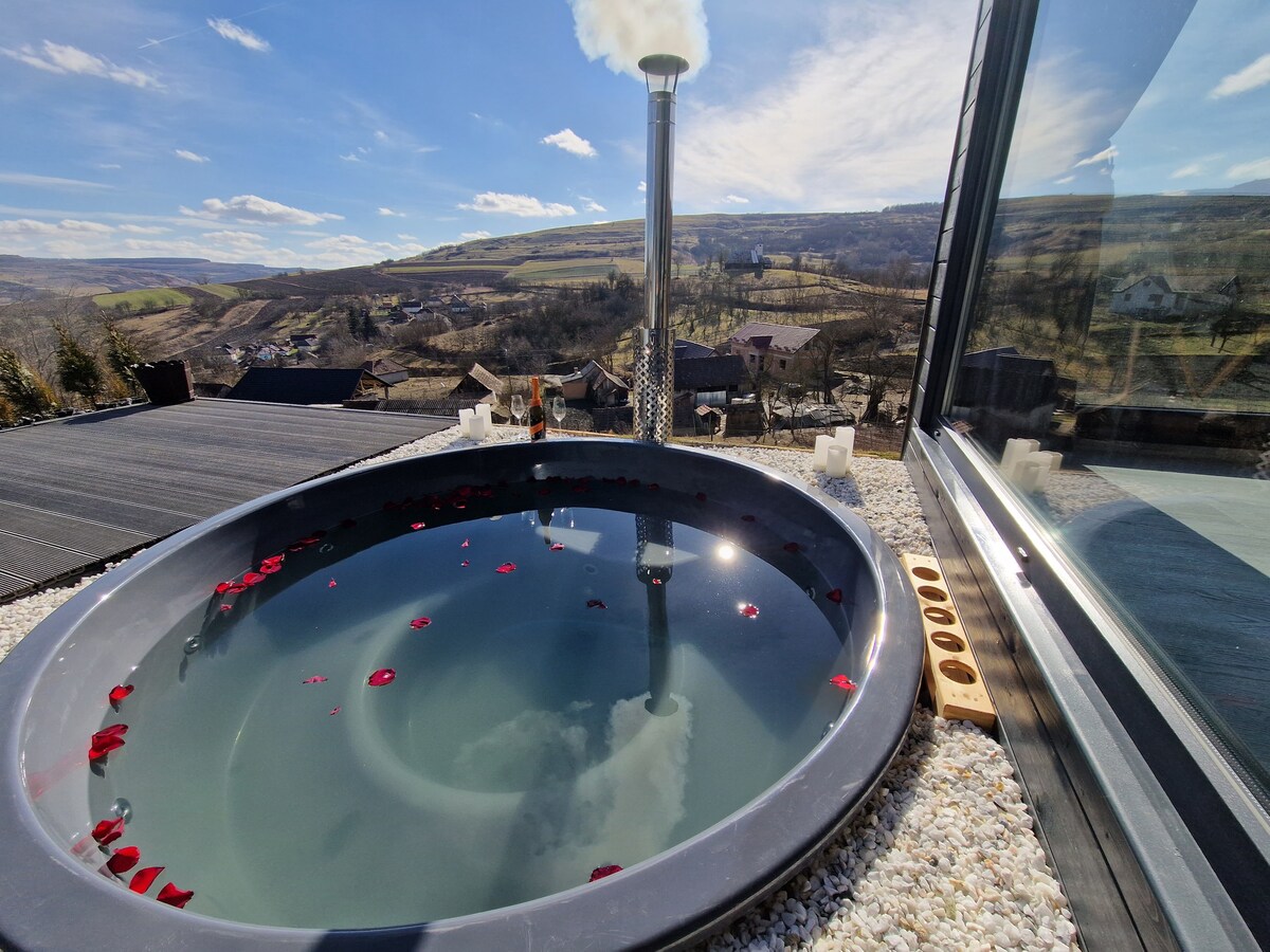 A circular hot tub is filled with water reflecting clouds and the sky, situated on a balcony overlooking rolling hills and a tranquil landscape. Scattered rose petals float on the surface, and a chimney is visible nearby, complementing the serene outdoor setting.