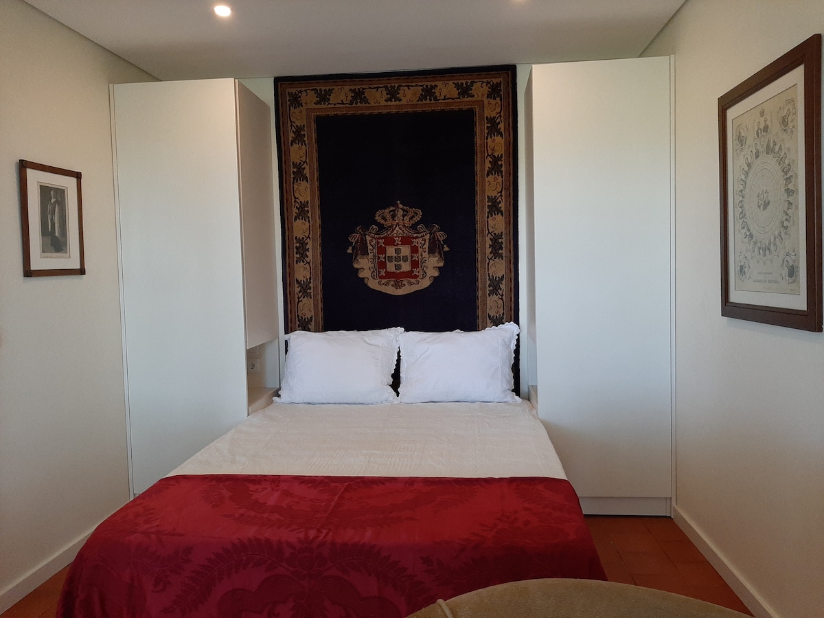 A cozy sleeping area features a spacious bed with white linens, complemented by a red bedspread. A decorative wall tapestry hangs above the bed. Two built-in wardrobes flank the sides, along with framed wall art providing a touch of character to the room.