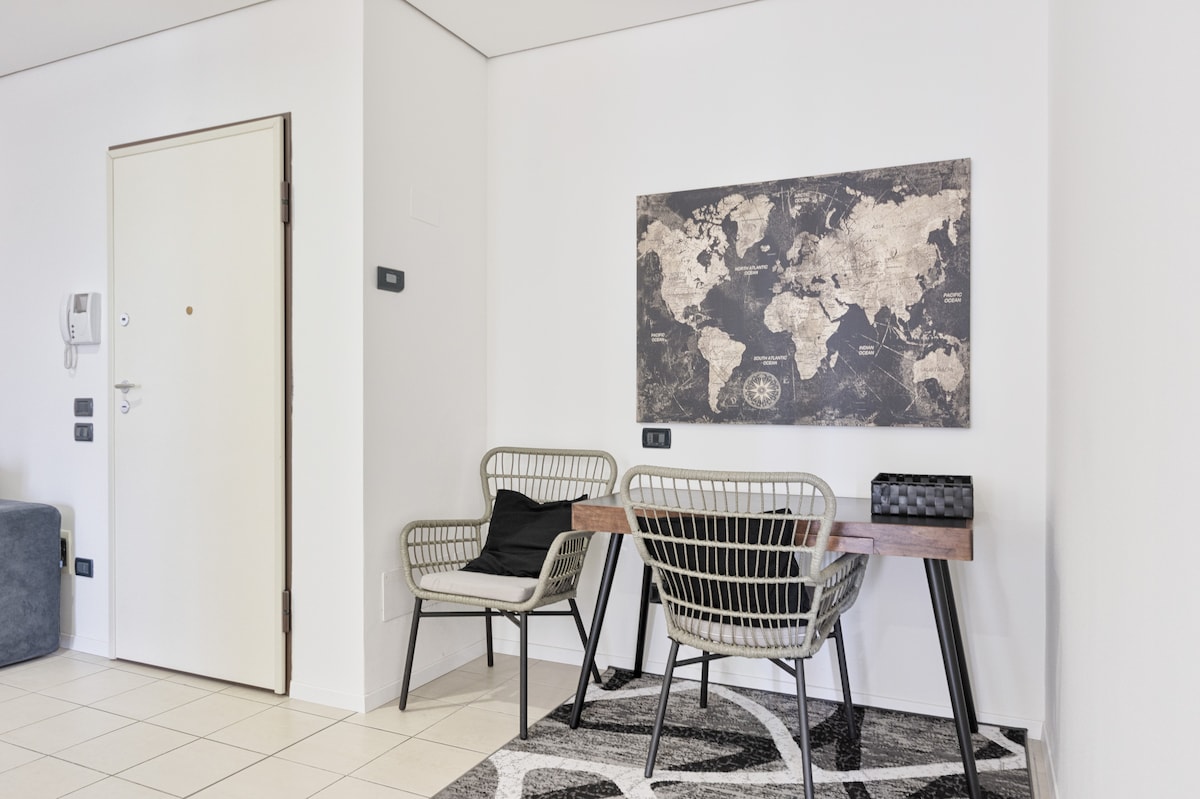 A modern area is featured with a wooden desk and two wire chairs, accompanied by a black storage box. A large world map is displayed on the wall, adding a global touch. Light tile flooring and a patterned rug complete the space.
