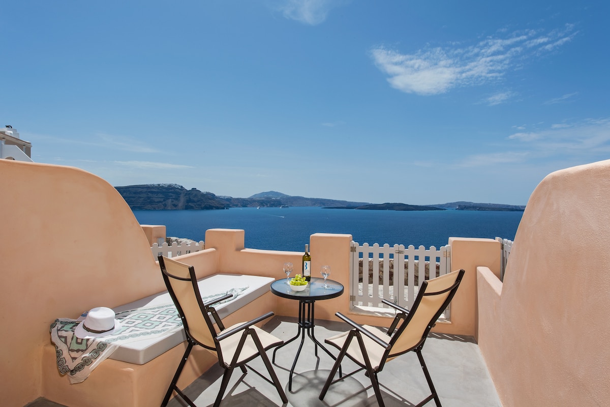 A sunlit terrace features a small round table with two glasses and a bottle, surrounded by two comfortable chairs and a lounging area. The panoramic view displays the vibrant blue sea, with gentle waves and a clear sky overhead.