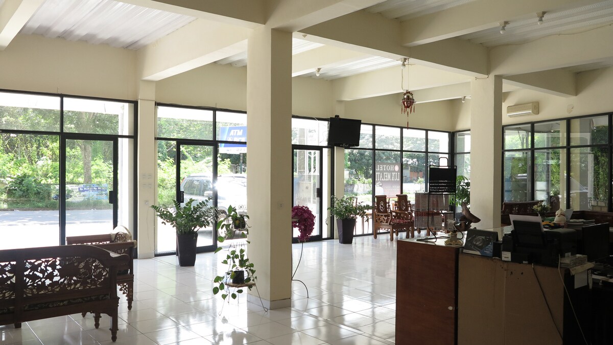 A spacious lobby with floor-to-ceiling windows allows natural light to fill the area. Wooden furniture is arranged for seating, complemented by decorative plants. A reception desk is visible to one side, providing a welcoming entrance for guests.