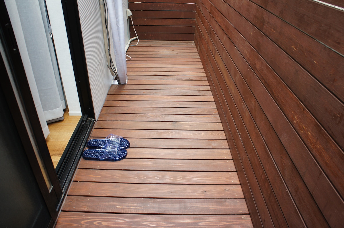 A small wooden deck is shown, featuring smooth, polished planks. A pair of blue slippers is placed casually at the edge near the entrance. The surrounding wooden walls provide a cozy and enclosed feel to the space.