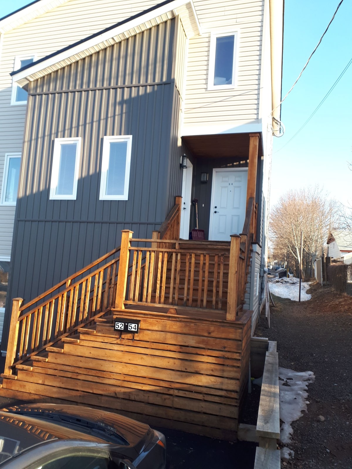 The exterior of a two-story loft apartment is shown, featuring a wooden staircase leading to a covered entrance. The building displays a mix of gray and light-colored siding. Snow is visible at the base of the steps, indicating the season.