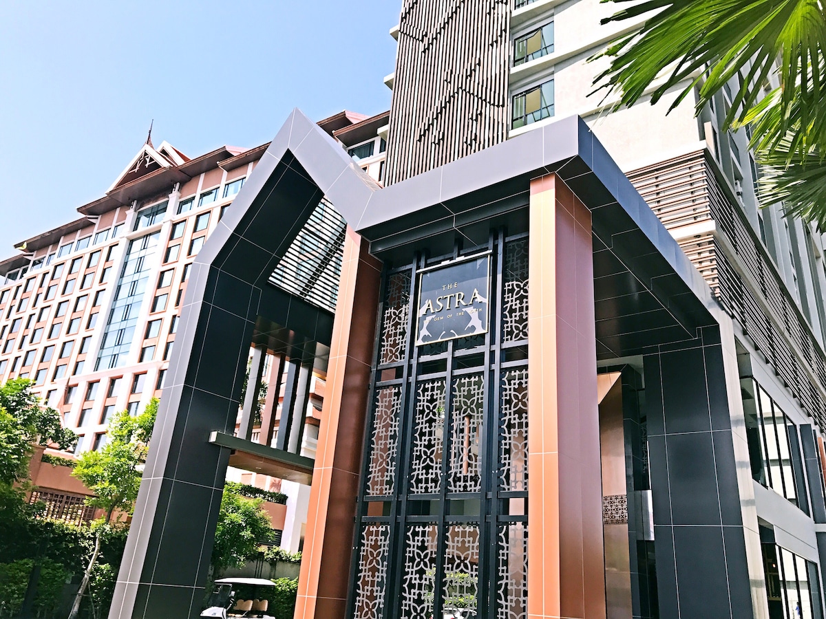 The entrance of the Astra condominium complex is highlighted by modern architecture, featuring a sleek façade with glass and metal accents. Lush greenery surrounds the entrance, enhancing the inviting atmosphere. The building's name is prominently displayed above the entryway.