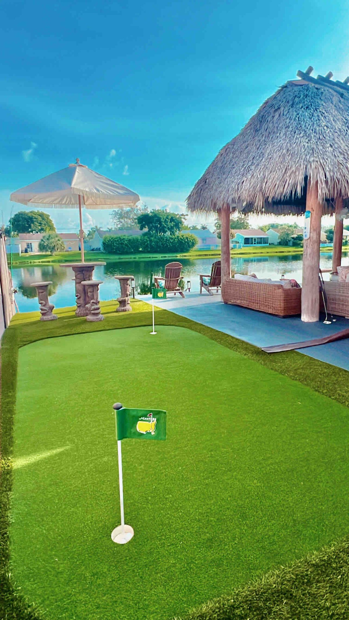 A private golf putting green is set on well-maintained artificial turf, accompanied by a shaded tiki hut and outdoor seating. A white umbrella provides shade, while a serene lake reflects the blue sky, enhancing the peaceful ambiance of the backyard.