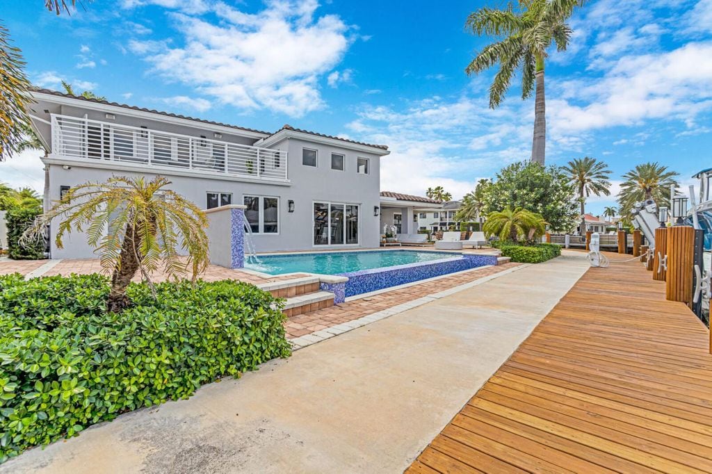 An expansive outdoor area features a large pool surrounded by tropical landscaping. Palm trees stand tall near the edge of the pool, while a wooden deck extends toward the dock area. The striking blue of the pool contrasts with the neutral tones of the house.