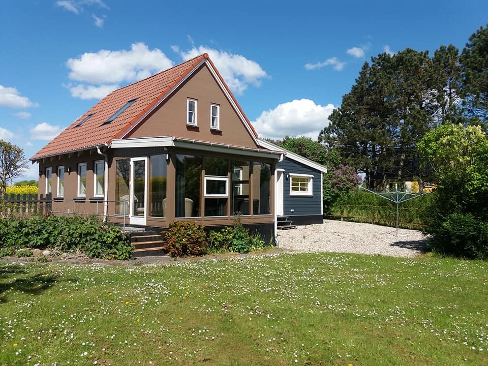 The house features a two-story design, with large windows allowing natural light to fill the interior. The surrounding garden showcases a grassy lawn adorned with daisies, complemented by mature trees in the background. A clothesline is visible to the side, enhancing the outdoor space.