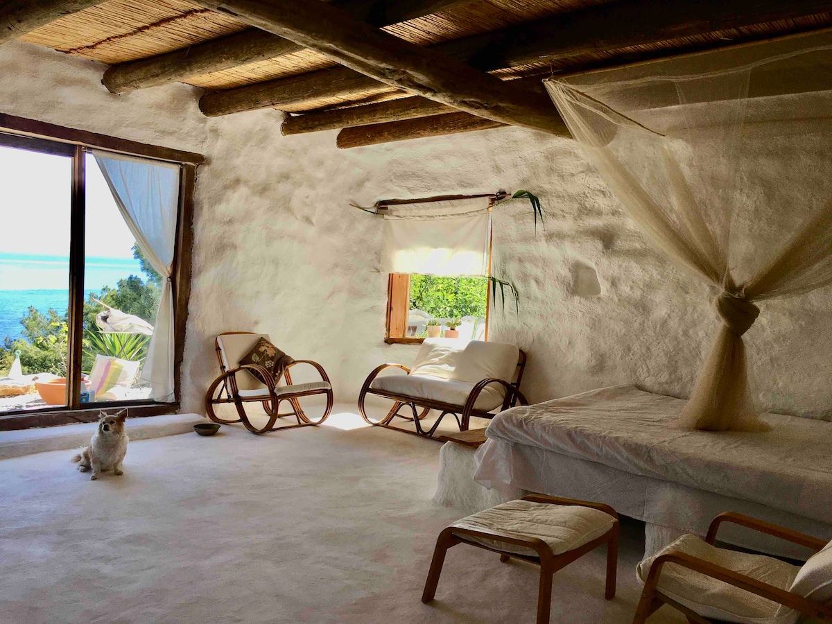 A bright interior space features a spacious bed draped in a light canopy, complemented by wooden beams overhead. Two rocking chairs and a small table are arranged near an expansive window, offering views of the picturesque landscape beyond. A small dog sits calmly on the floor.