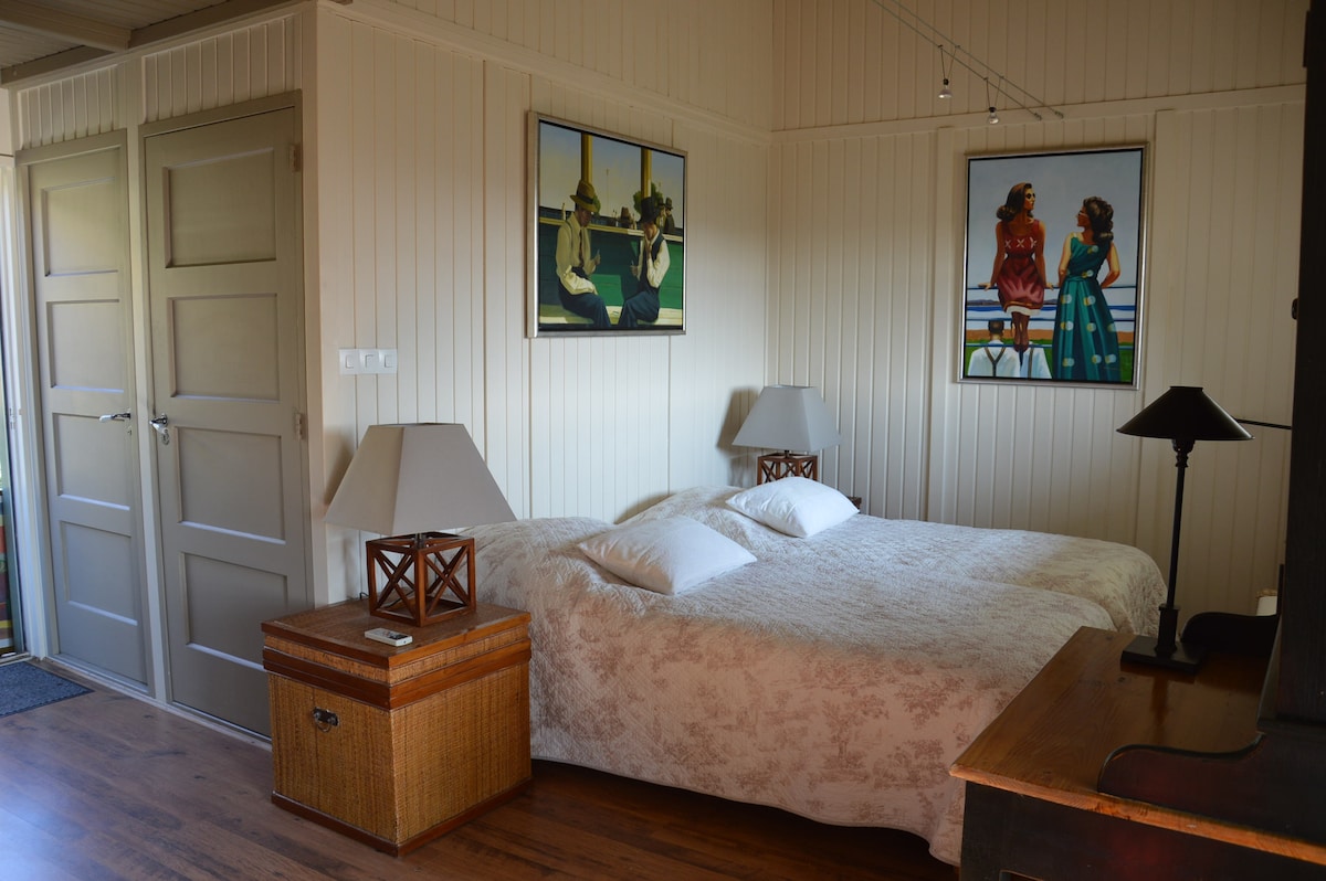 A well-lit sleeping area features two beds with white linens, positioned next to wooden nightstands with lamps. Artwork adorns the walls, depicting figures in serene settings. A wicker chest and wooden desk add practicality to the room. The space is complemented by natural light from a nearby entrance.