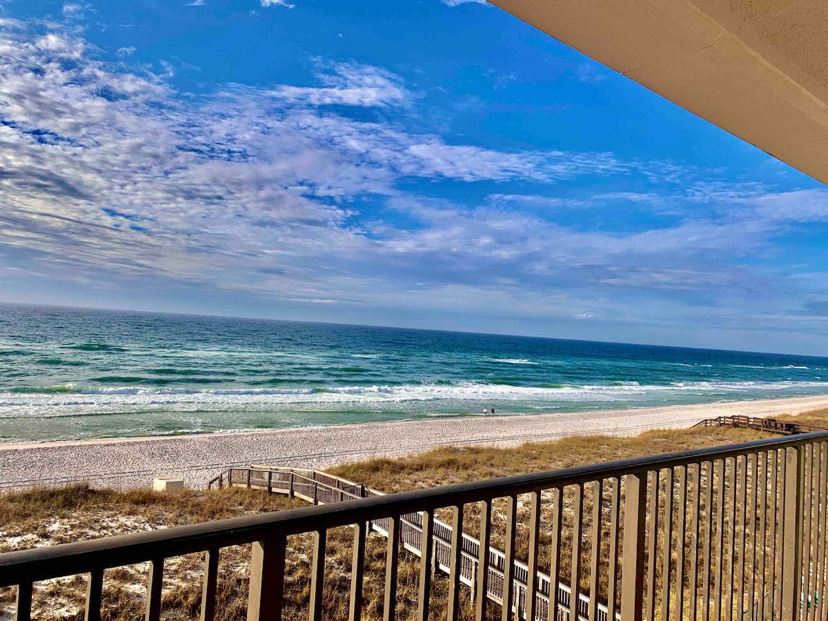 A view of the beach and ocean is presented from the balcony. Gentle waves can be seen rolling onto the shore, alongside a wide stretch of sand. The sky features a mix of clouds and blue tones, creating a serene coastal atmosphere.