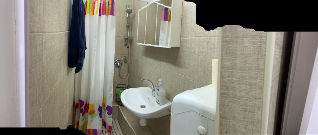 The bathroom features a compact layout with light-colored tile walls. A shower area is equipped with a modern showerhead, and a white sink is mounted beside a mirror. A decorative shower curtain adds a pop of color, complementing the overall neutral tones.